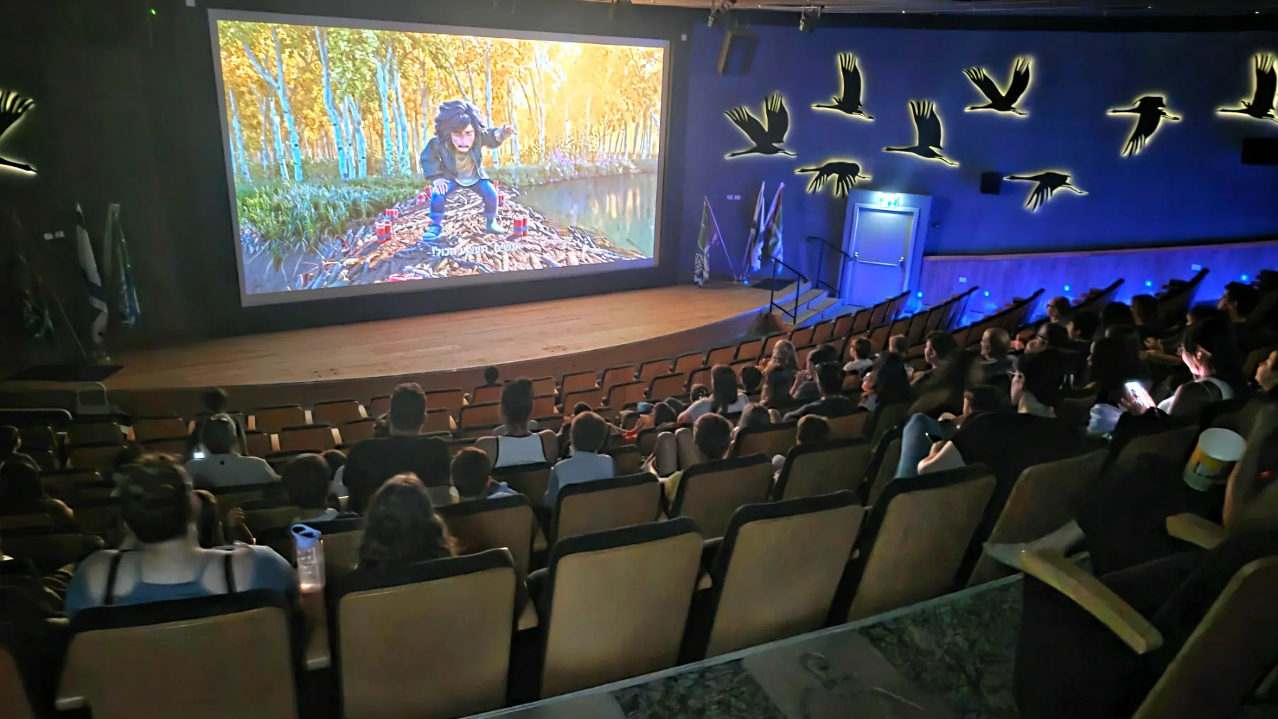 קהל תלמידים בהקרנת סרט אנימציה באודיטוריום, על רקע העגורים המוארים